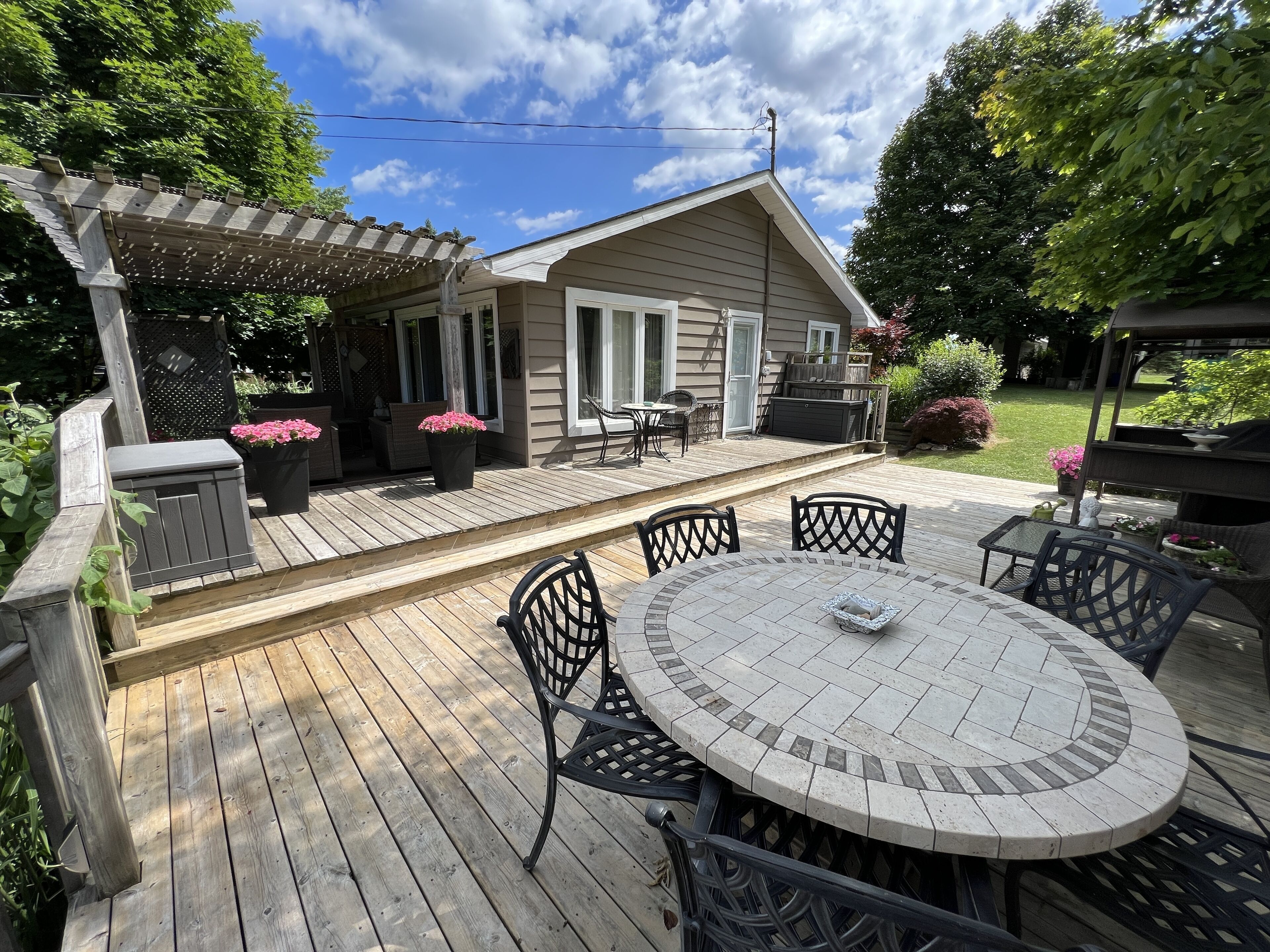 Een houten terras met een ronde tafel en stoelen, een pergola en een huis met een overdekte patio.