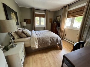 Quarto com cama, mesa de cabeceira, luminária, cômoda e janelas com cortinas.