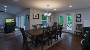 A dining area with a large wooden table and chairs, a television on the left, and a view of greenery through the glass doors. - Top Location! 3D & Video Tour. Riverside 4/3 w/ the best water access & privacy! (Flippin)