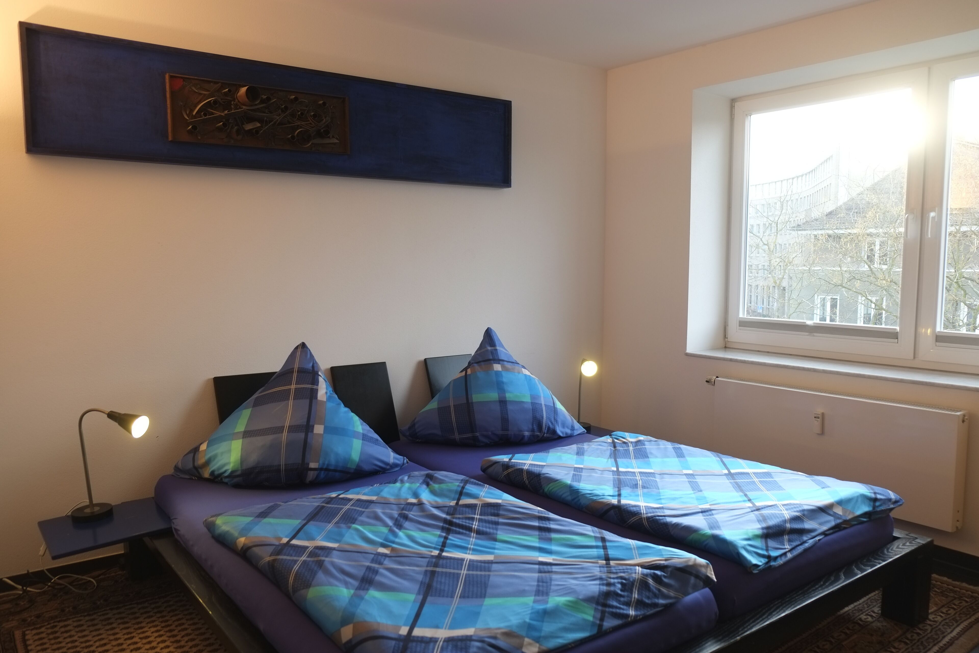 A room with two beds, a nightstand, a wall-mounted artwork, and a window with a view of buildings.