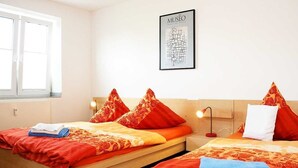A hotel room with two beds, each with red and orange bedding, a framed map on the wall, and a window providing natural light. - Nice, centrally located apartments in Cologne, streetcar stop in front of the door. (Köln)
