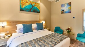 Una habitación de hotel con una cama grande, dos sillas azules, una mesita con un jarrón de flores y dos cuadros en la pared.