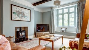 A cozy living room with a fireplace, a television, and a large window with curtains. - The Bladon Farmhouse | Oxfordshire Holiday Let | 2 Beds & Sleeps 5 (Bladon)