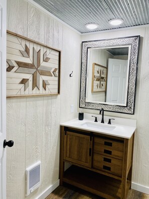 A bathroom with a white vanity, wooden cabinets, a large mirror with a decorative frame, and a geometric wall art piece. - TheBarnhouse: Secluded•Ride2Trails•PetFriendly•GameRoom• MountainRetreat•FirePit (Mena)