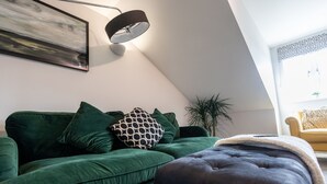 A green sofa with patterned pillows, a dark ottoman, and a wall-mounted lamp.