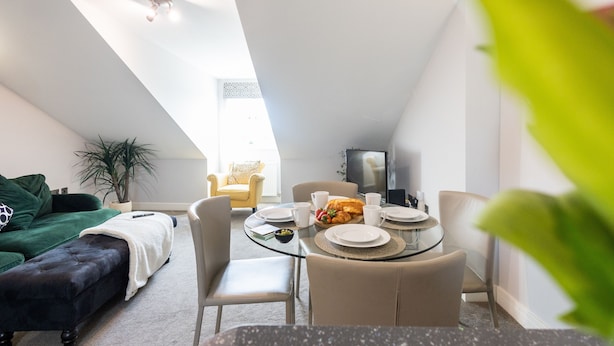 A modern living room with a dining area, featuring a round glass table, beige chairs, a dark sofa, and a green armchair.