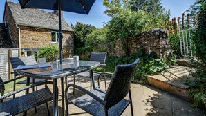 A patio with a glass table and wicker chairs, a stone building in the background, and a white fence with red flowers. - Rose Cottage | Adderbury Holiday Let | 2 Beds & Sleeps 3 (Adderbury)