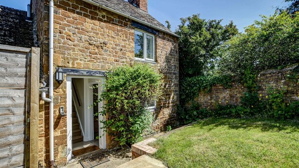 A stone house with a wooden door, a small front yard, and a brick wall. - Rose Cottage | Adderbury Holiday Let | 2 Beds & Sleeps 3 (Adderbury)