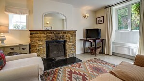 A living room with a stone fireplace, a television, and a large window with curtains. - Rose Cottage | Adderbury Holiday Let | 2 Beds & Sleeps 3 (Adderbury)