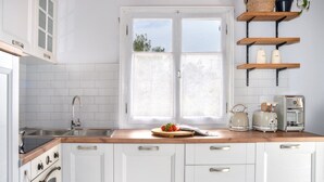 Una cocina con armarios blancos, encimera de madera, ventana con cortinas de encaje y electrodomésticos antiguos.