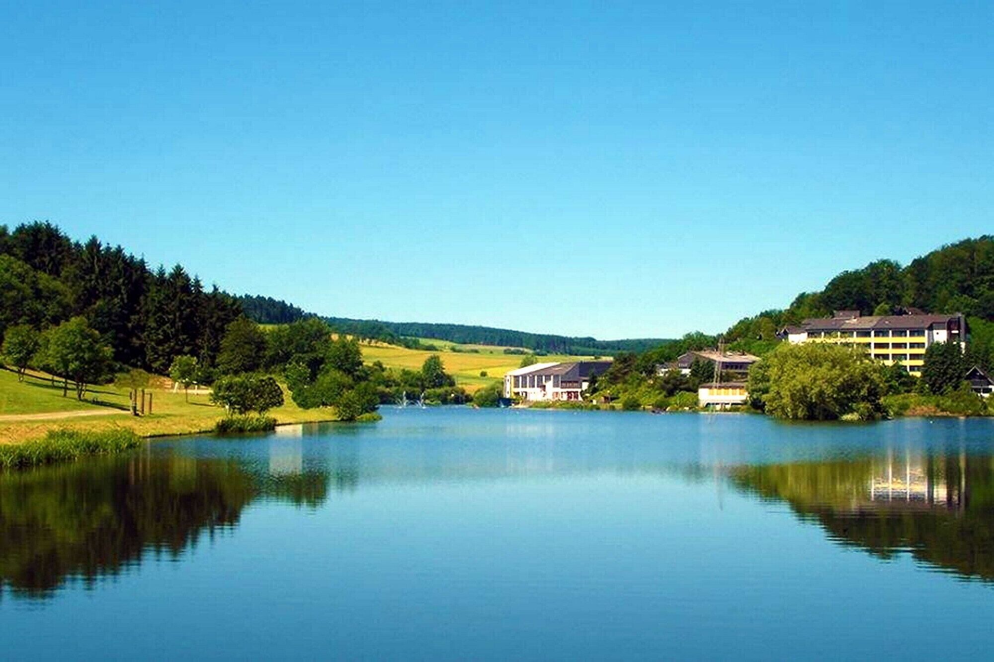 Ein stiller See mit Gebäuden rechts und einem bewaldeten Hügel links.