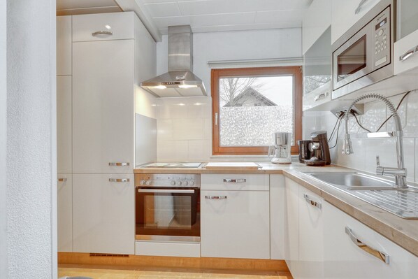 A modern kitchen with white cabinetry, a built-in oven, a stainless steel range hood, a window with a view, and a coffee maker. - Vacation home near Viola (own sauna, 50''-TV, WLAN) in the forest, by the lake (Kirchheim)