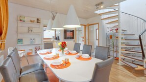 Un comedor moderno con una mesa blanca para cuatro personas, rodeada de sillas grises. Se ve una escalera a la derecha, con peldaños de madera y barandillas de metal.
