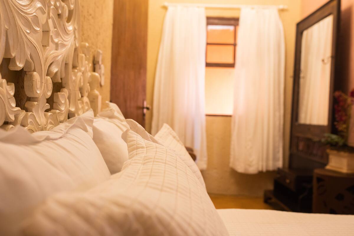 A bed with ornate headboard, white curtains, and a mirror on a bedside table.