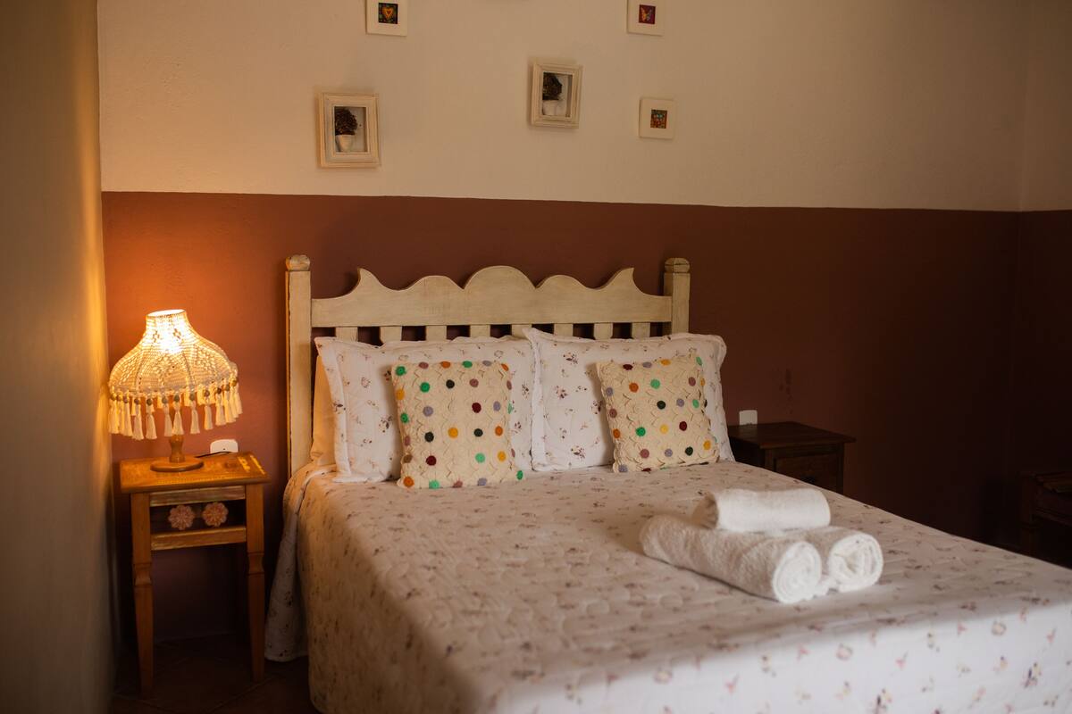 A neatly made bed with a floral patterned bedspread, a wooden headboard, and two pillows with polka dots. A small bedside table with a lamp and a folded towel is visible.