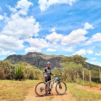 En mountainbiker står ved siden af en cykel på en jordvej med et bjergrigt baggrund og en klar himmel.
