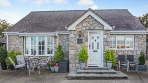 Ein Steinhaus mit weißer Tür, zwei Fenstern und einer kleinen Veranda mit Stühlen und Topfpflanzen.