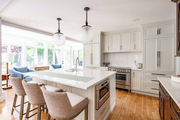 A modern kitchen with white cabinetry, a central island with bar stools, stainless steel appliances, and large windows offering a view of the outdoors.