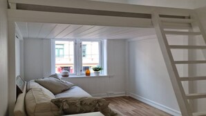 A room with a loft bed, a small table, a window with a view of buildings, and a wooden ladder. - Oslo International Designers Space (Oslo)