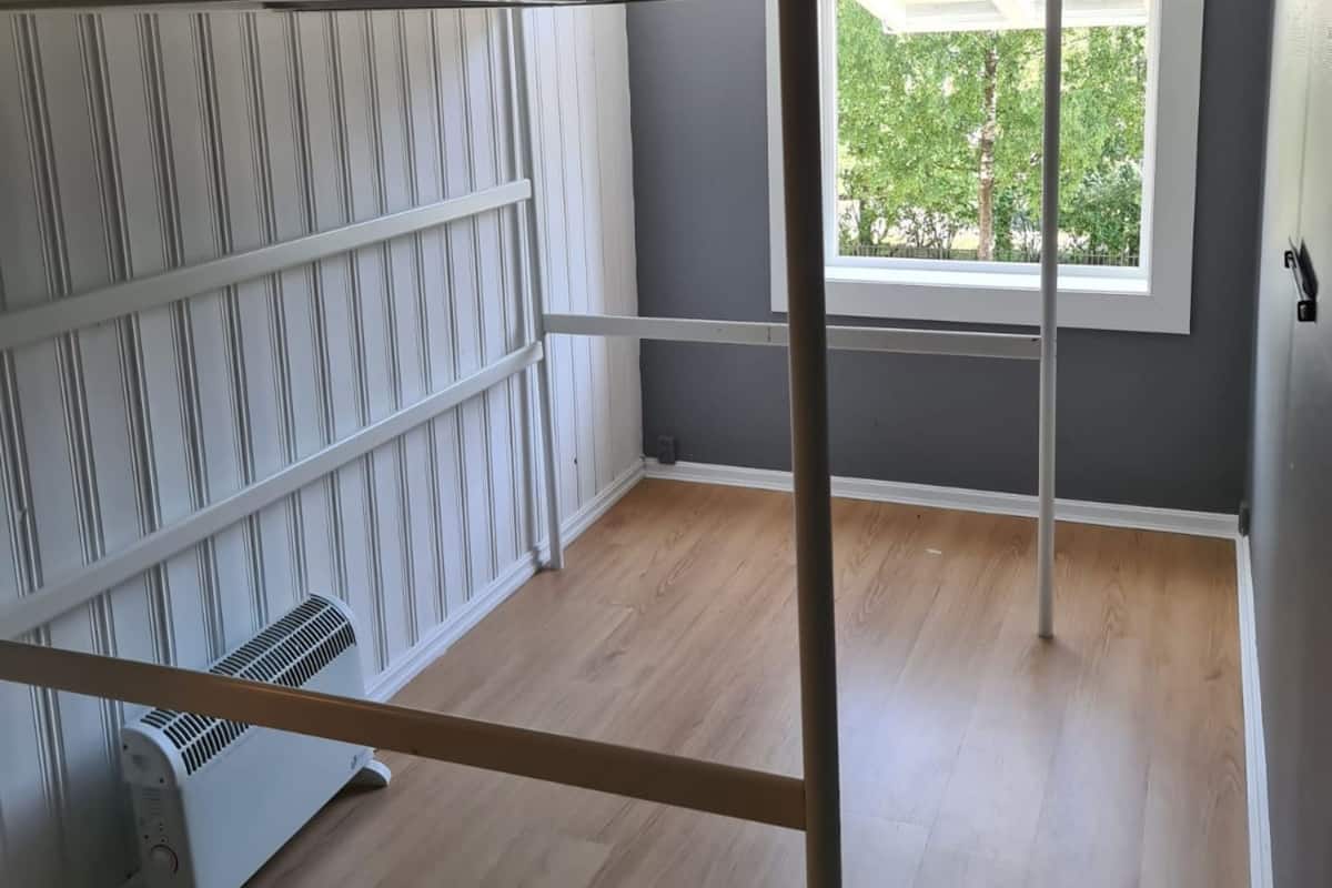 A room with a bunk bed, a radiator, a window with a view of trees, and a grey wall.