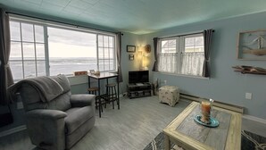 A living room with a sofa, a small table, a TV, and a view of the ocean through large windows. - Bayview #1 on Lake Huron (Saint Ignace)