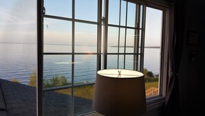 A room with a large window overlooking a body of water, a hanging lamp, and a view of a grassy area outside. - Bayview #1 on Lake Huron (Saint Ignace)