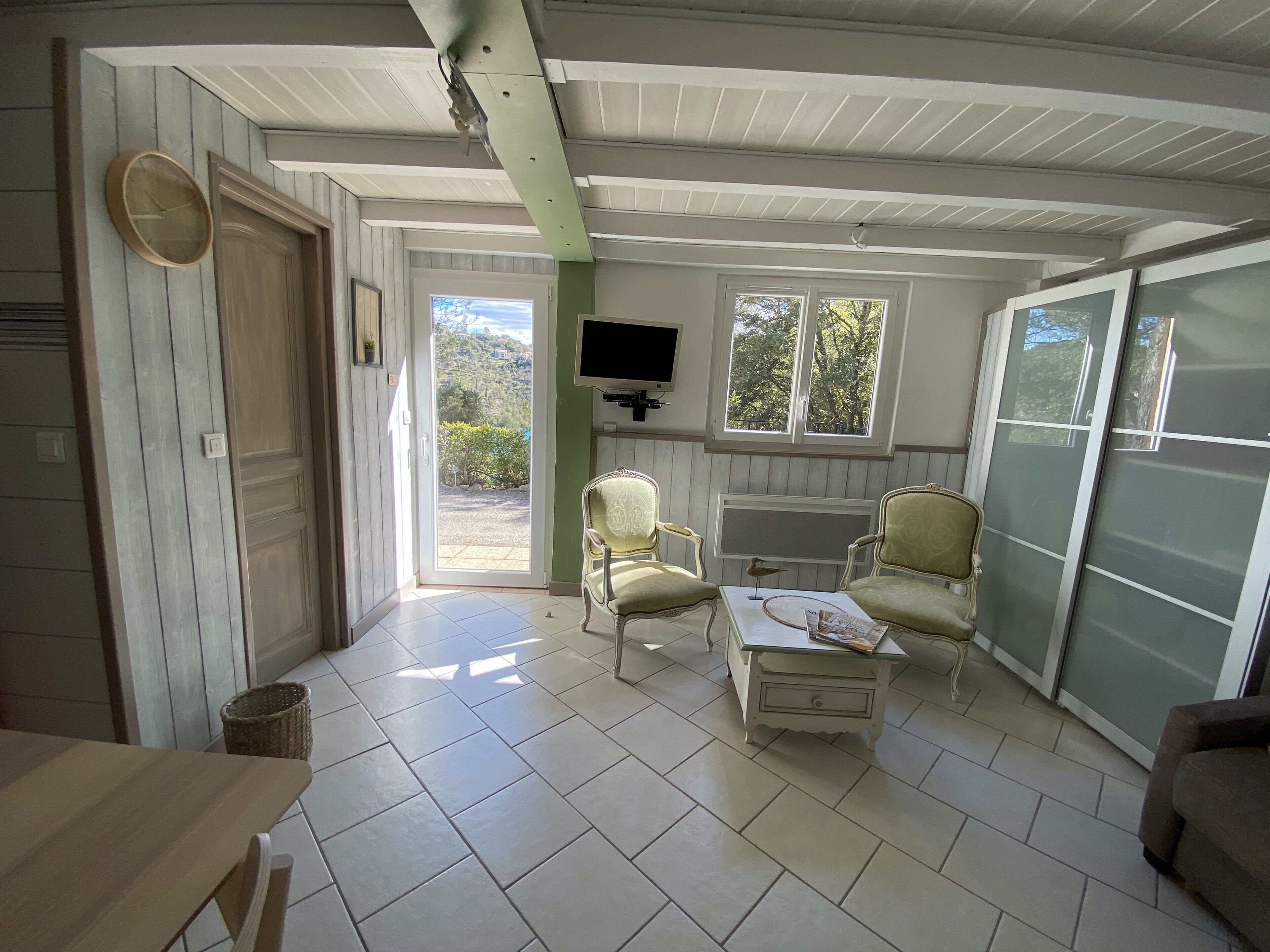 A room with a large sliding glass door leading to a balcony, two armchairs, a small table, and a television mounted on the wall.
