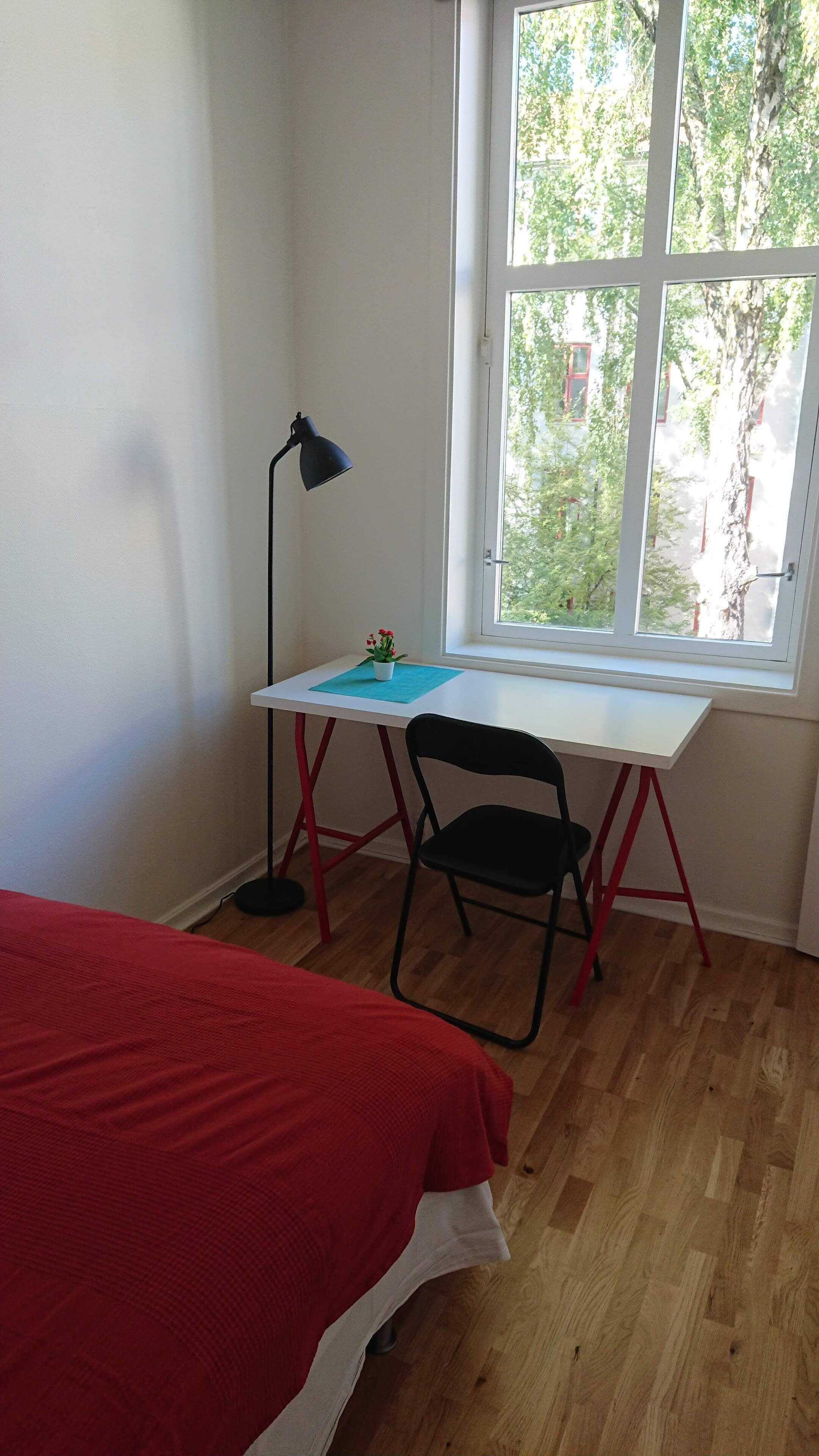 Een kamer met een bed, een bureau met stoel, een lamp en een raam met uitzicht op bomen.
