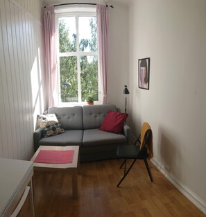 Un salon avec un canapé gris, un coussin rouge, une petite table, un lampadaire et une chaise. Il y a une fenêtre avec des rideaux, une plante sur le rebord de la fenêtre et un tableau encadré au mur.