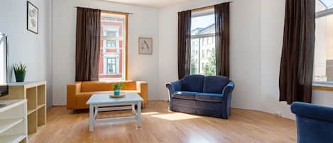 A living room with a wooden floor, a white coffee table, a blue sofa, and a brown sofa. There are two windows with brown curtains, one with a view of buildings and another with a view of trees.