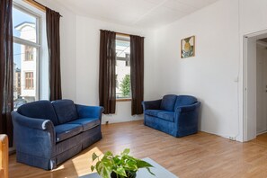 A room with two blue sofas, a wooden floor, a white wall with a pineapple painting, and a window with brown curtains.