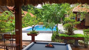 A pool area with a thatched roof, surrounded by lush greenery and traditional buildings. - Tam Coc Melody Homestay (Nam Hoa Lu)