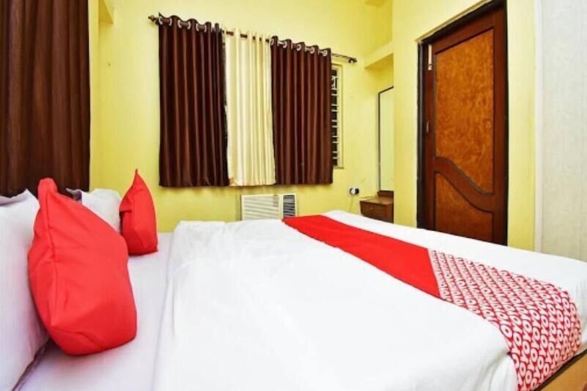 Quarto de hotel com cama, almofadas vermelhas, manta com padrão vermelho e branco, porta de madeira e cortinas.