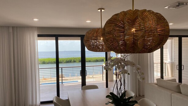 Un comedor moderno con una mesa de madera, sillas blancas, una planta en maceta y vistas al mar a través de amplios ventanales.