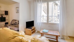 A living room with a sofa, a television on a wooden stand, a coffee table with a book, and a window with a balcony view.