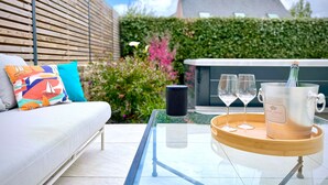 Un espace extérieur moderne, aménagé avec une table en verre, un canapé blanc agrémenté de coussins colorés et un réfrigérateur à vin avec des verres.