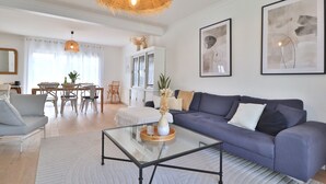 Un salon moderne avec une table basse en verre, un canapé bleu et un coin repas comprenant un buffet blanc et un lustre.