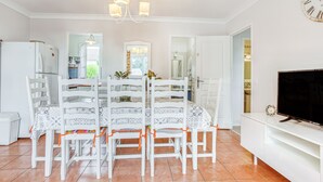 A dining area with white chairs and a table, a white refrigerator, a TV on a white stand, and a clock on the wall.