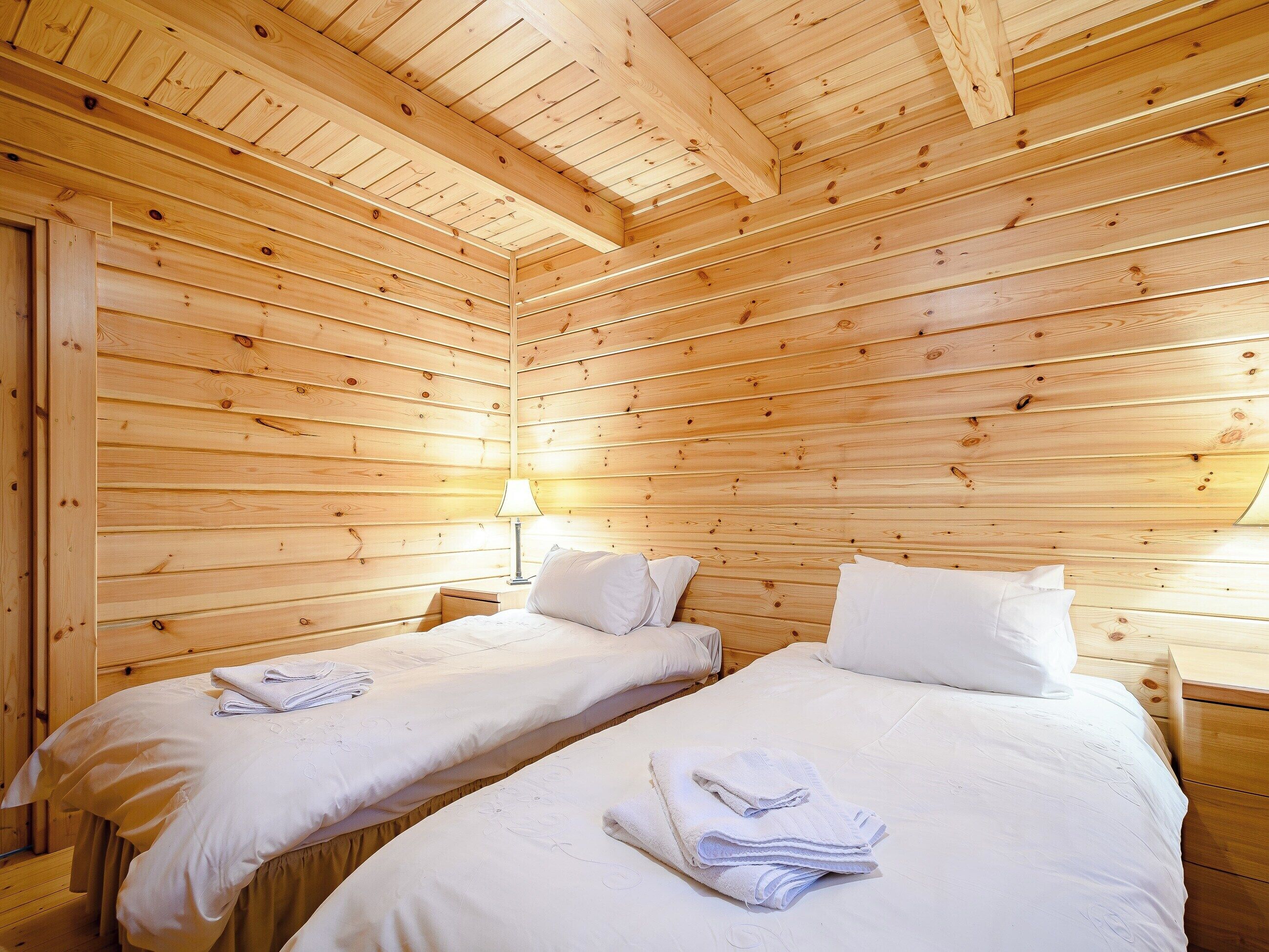 A wooden cabin room with two beds, a nightstand, and a lamp.