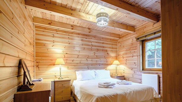 A wooden cabin bedroom with a bed, two bedside tables with lamps, a radiator, and a window. - 2 bedroom accommodation in Killin, near Crianlarich (Killin, near Crianlarich)