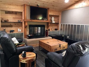 Un salon chaleureux avec une cheminée, des canapés en cuir, une table basse en bois et un téléviseur fixé au-dessus de la cheminée.