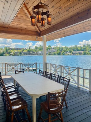 A covered deck with a table and chairs, overlooking a lake with buildings in the background.