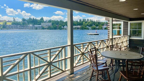 En inglasad veranda med utsikt över en sjö, en brygga med en person på och ett bord med stolar.
