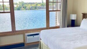 A hotel room with a large window offering a view of a body of water and greenery. The room has a bed with a white comforter and a nightstand with a lamp. - Lake Placid Club Boat House Residences 3 Bedroom (Lake Placid)