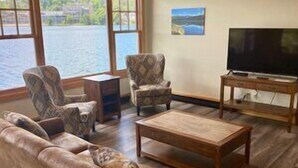 A living room with a large window, a sofa, armchairs, a coffee table, and a TV on a wooden stand. - Lake Placid Club Boat House Residences 3 Bedroom (Lake Placid)