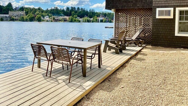 A wooden deck with a table and chairs overlooking a lake, a building with a dark brick exterior, and a clear sky. - Lake Placid Club Boat House Residences 3 Bedroom (Lake Placid)