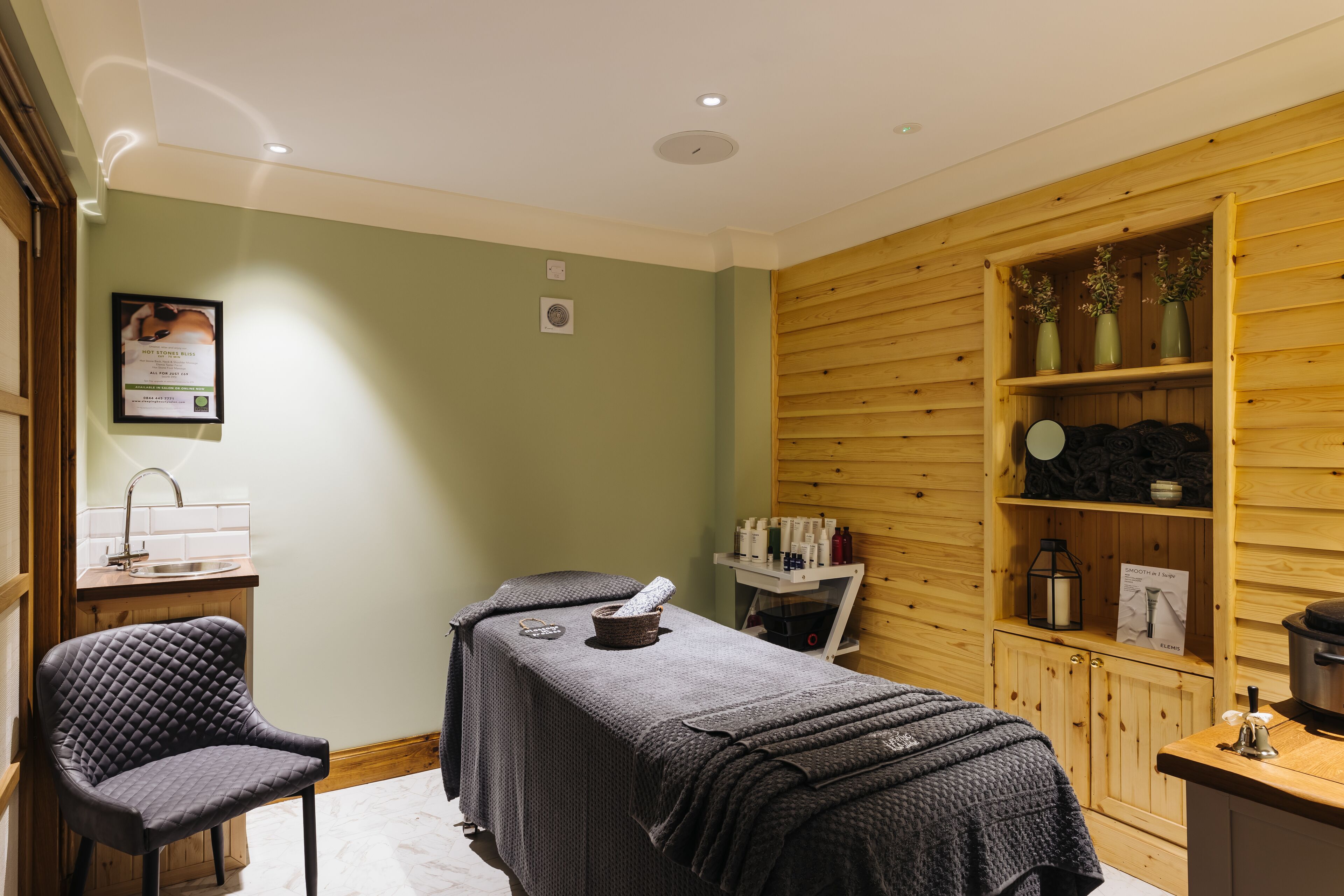 Een spa ruimte met een massagetafel, een stoel, houten lambrisering en een plank met flessen en een lantaarn.