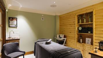 Een spa ruimte met een massagetafel, een stoel, houten lambrisering en een plank met flessen en een lantaarn.