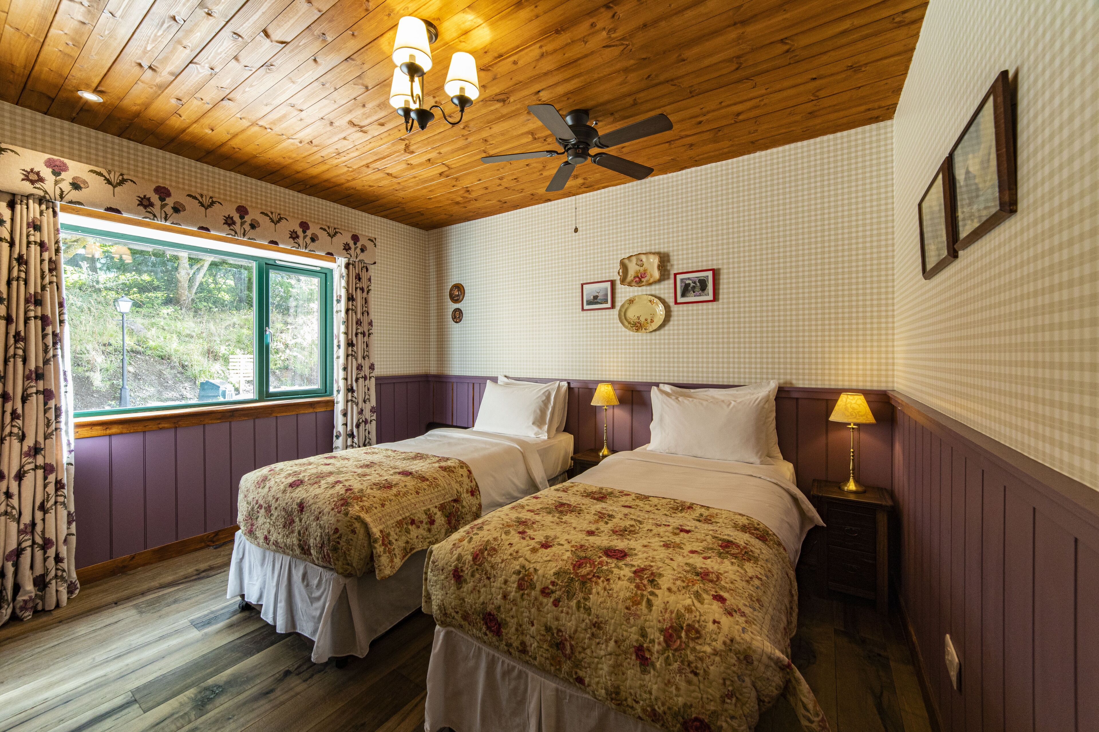 A room with two beds, wooden ceiling, floral curtains, and a window with a view of greenery.