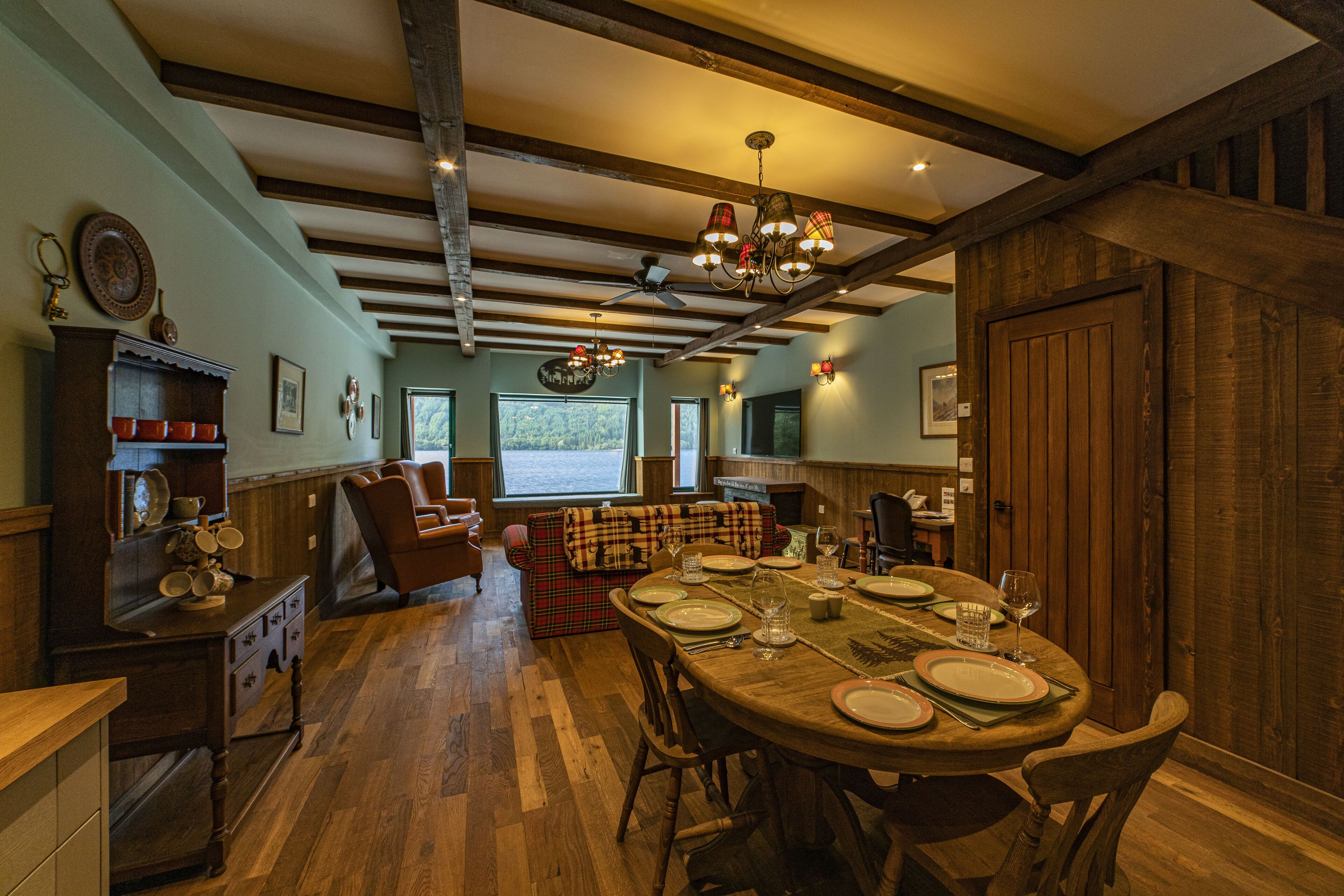 A dining area with a wooden table set for six, surrounded by wooden chairs. There is a sofa and a fireplace in the background.